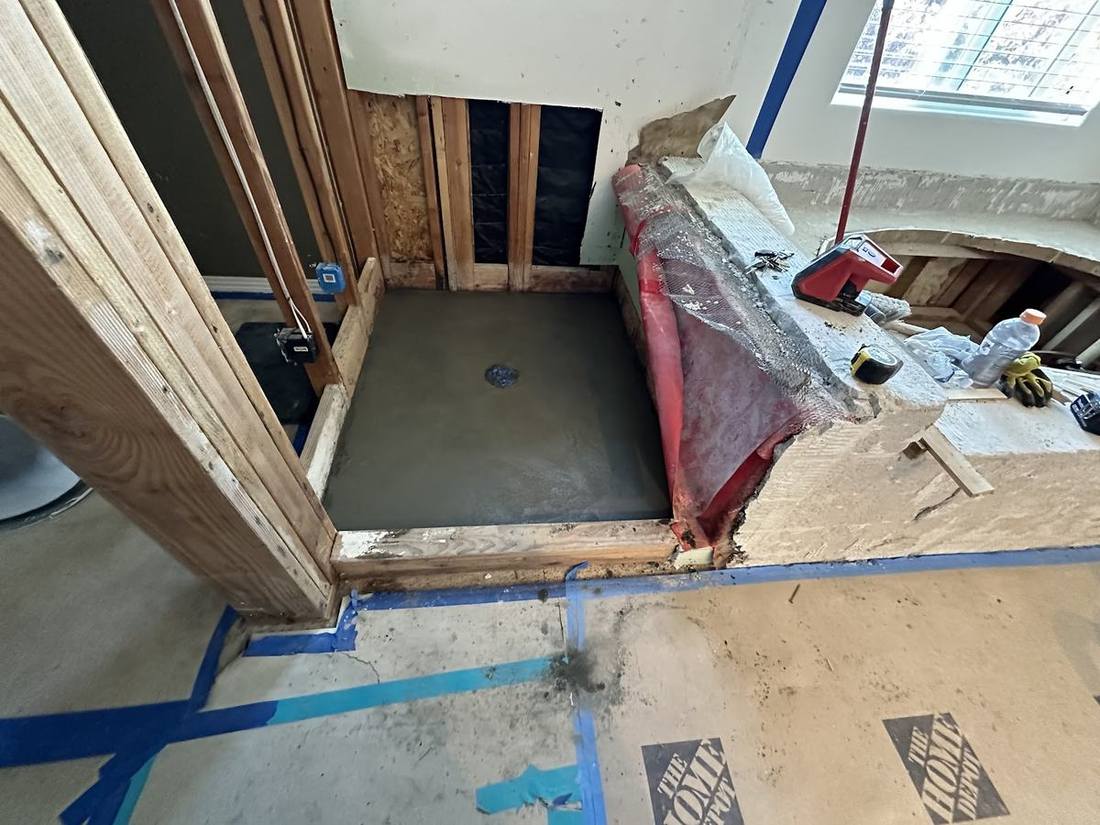 A bathroom at rough-in stage: a new shower pan, red waterproofing, framing and plumbing pipework in an open wall.