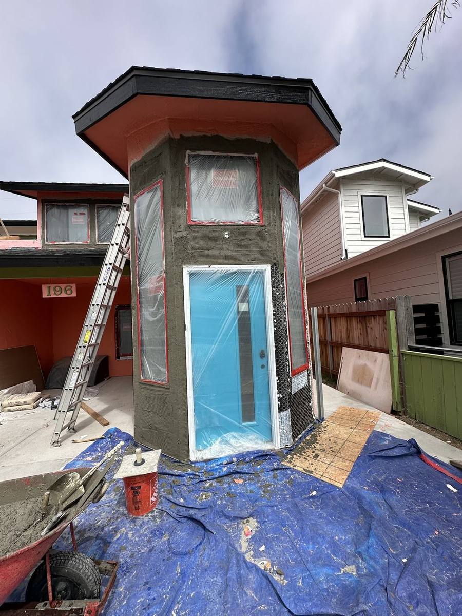 An exterior entry tower part-way through stucco work, with the door and windows masked in plastic and a ladder alongside.