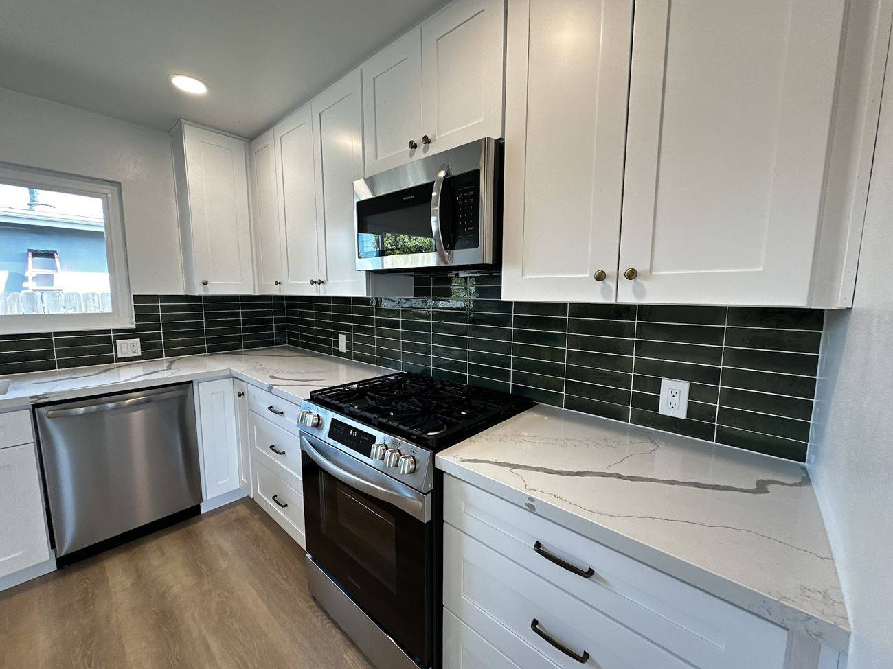 A finished kitchen with white shaker cabinets, a green tile backsplash, stone countertop and stainless appliances.
