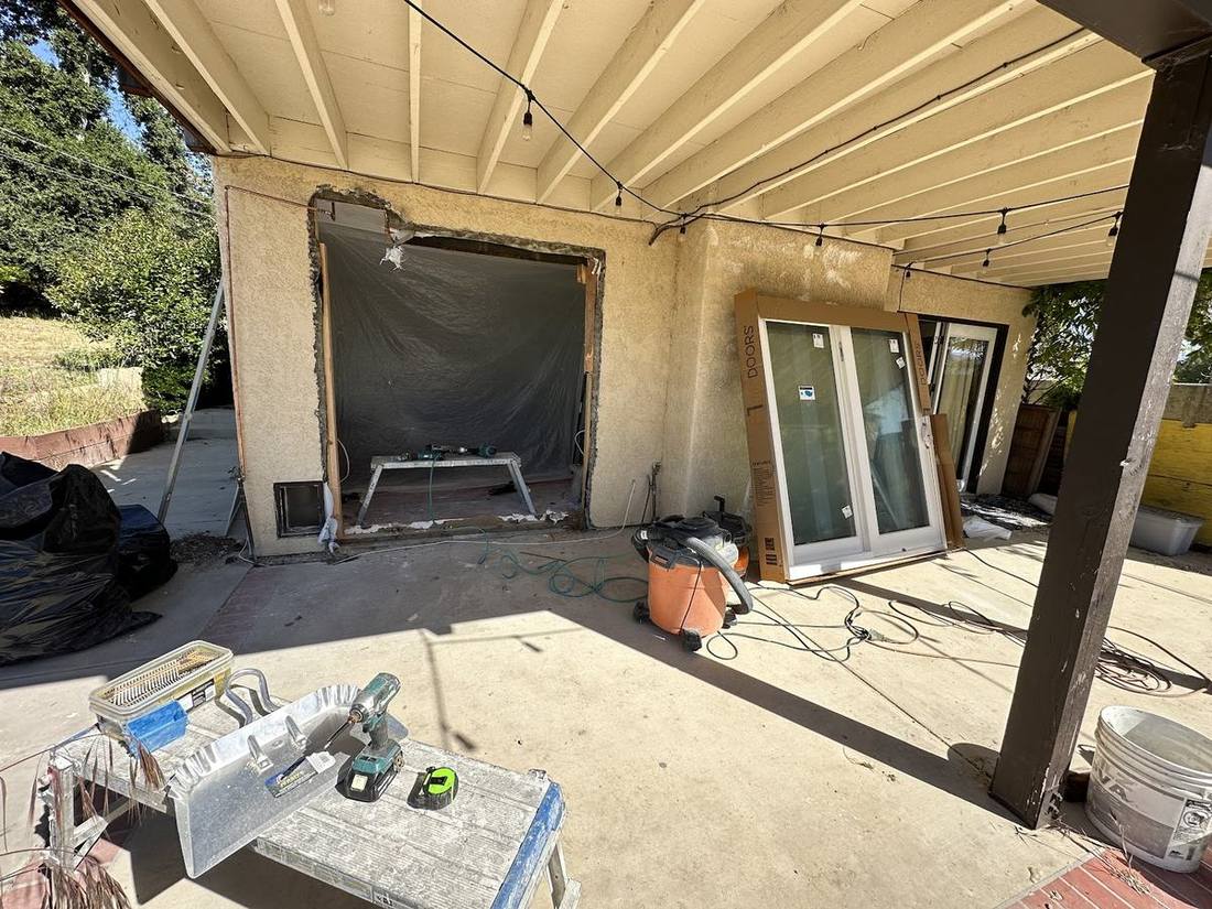 A covered patio with a large opening cut into the wall, sheeted over, and new sliding doors stacked ready to install.